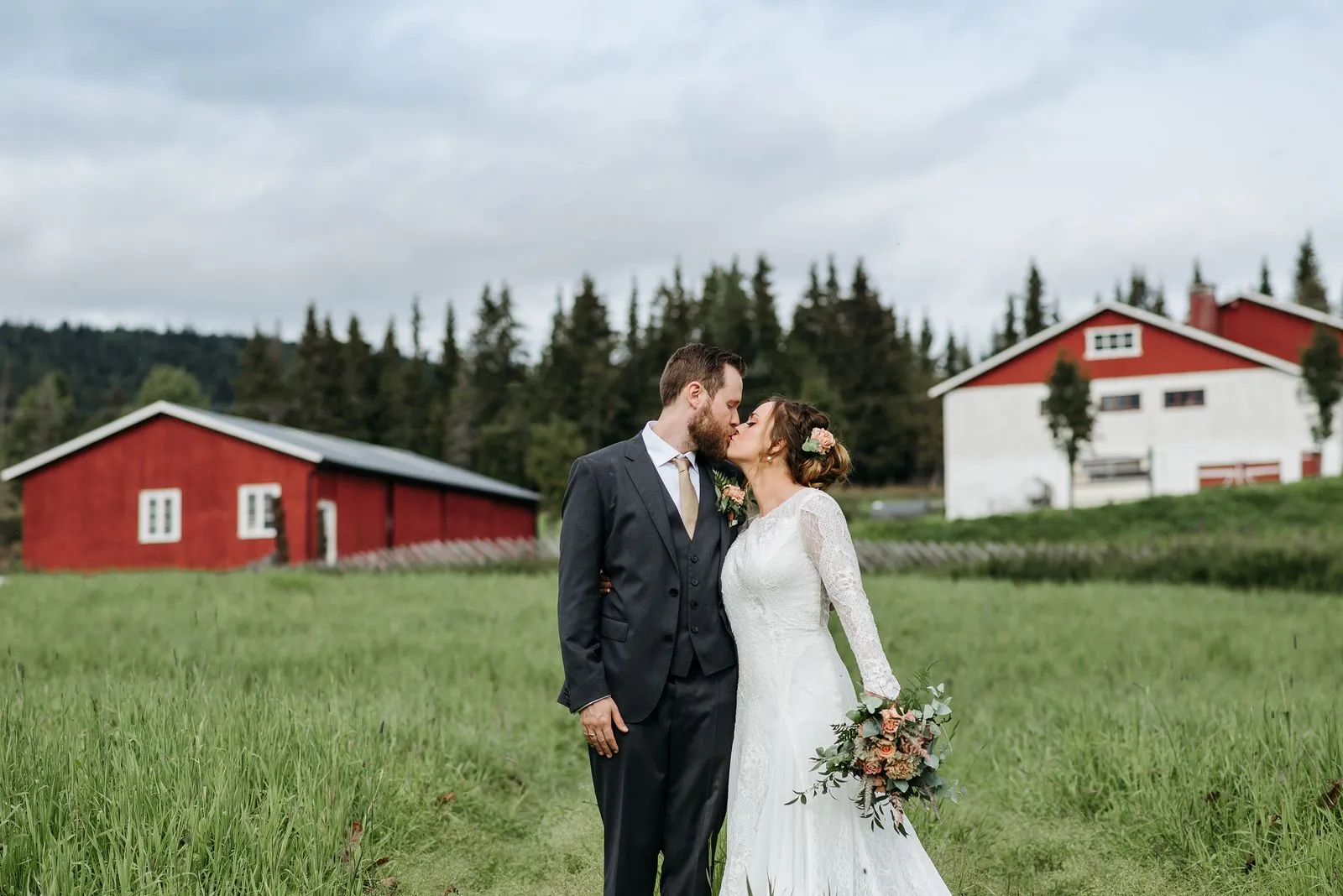 A bride and groom kissing in a green field in front of red farm buildings.