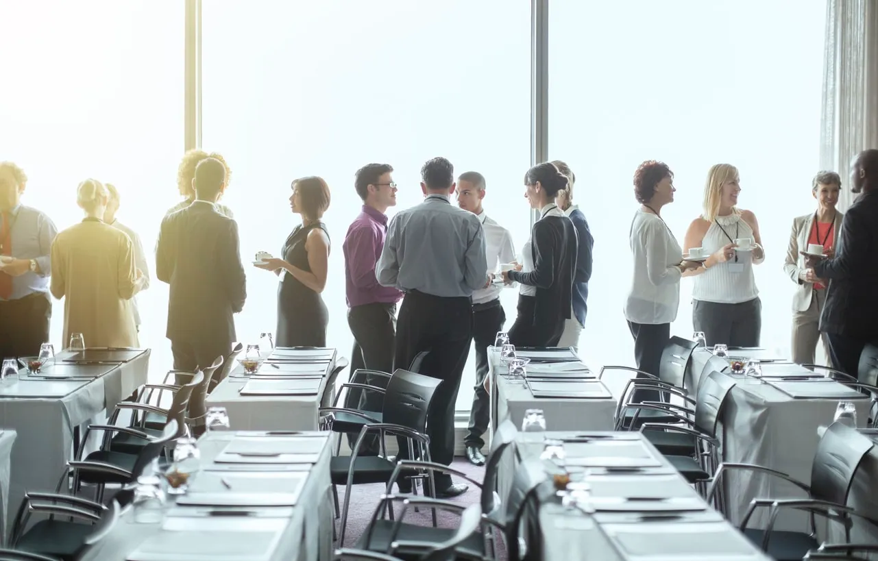 Event guests talking over coffee beside tall windows, with tables set for a meeting in the foreground.