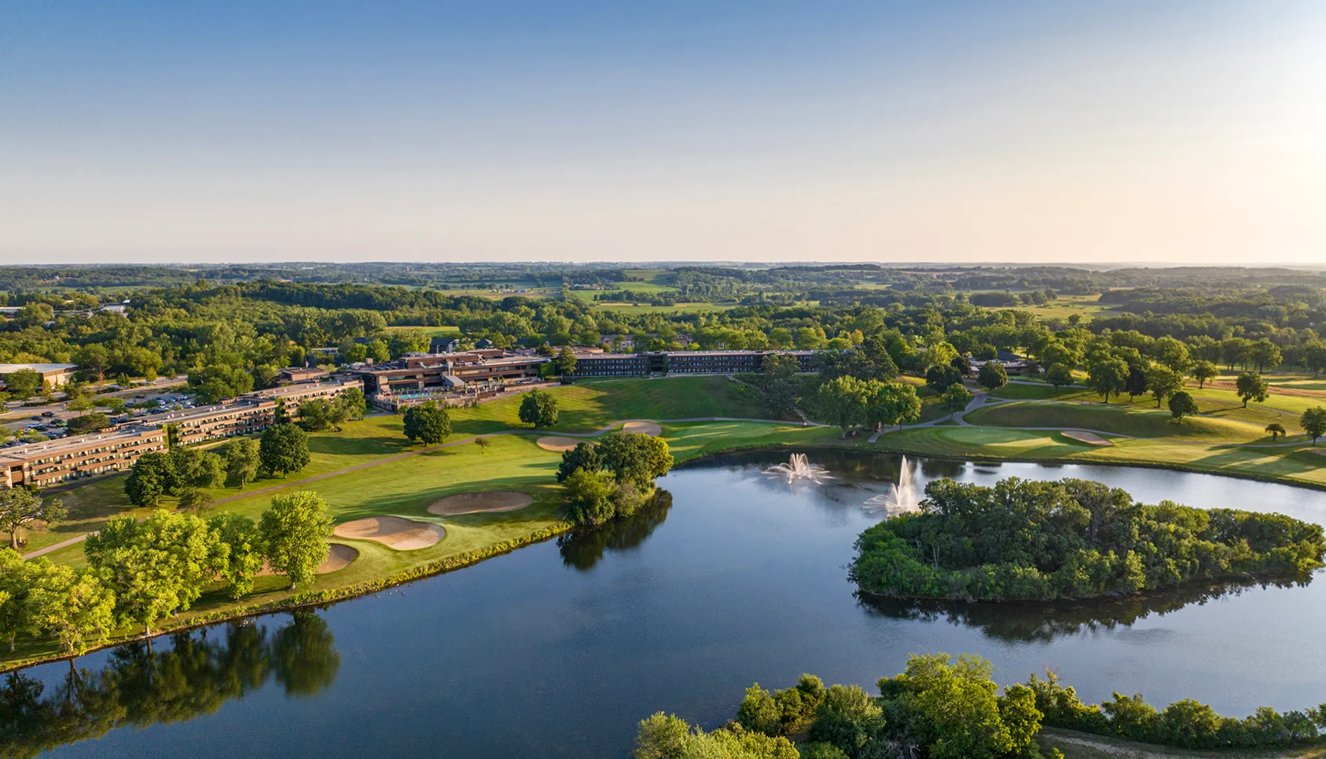 Aerial view of a resort beside a golf course and a pond with fountains, surrounded by rolling Wisconsin countryside.